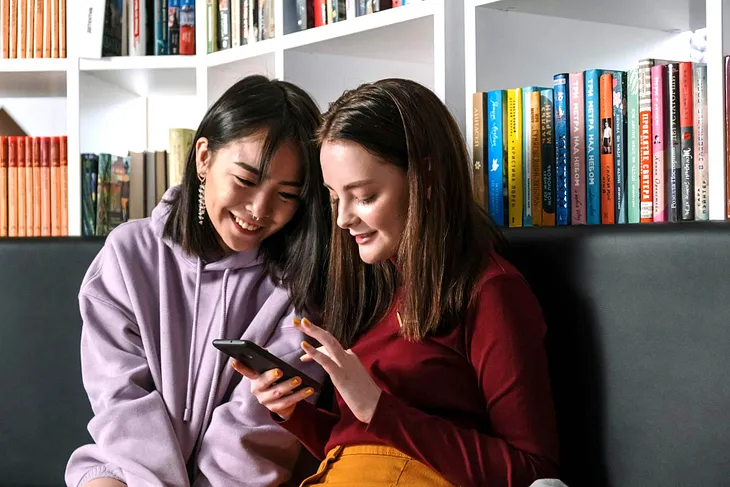 Two girls in front of bookshelves, talking, and looking at a phone