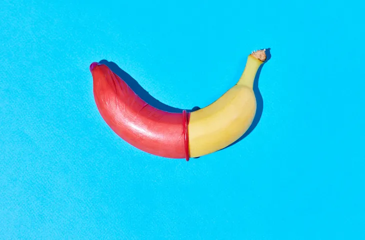 A banana lies on a bright blue background. The left half of the banana is covered with a red condom, creating a sharp contrast with the uncovered yellow right half. The image is brightly lit and has a playful, educational tone, likely symbolizing safe sex practices.