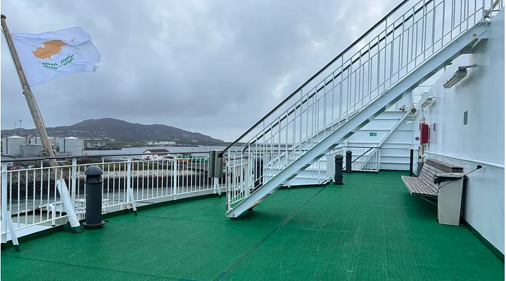 Deck of a Wales to Ireland ferry, with green astro turf, white railings and misty mountains in the distance. A flag whips in the wind on a stormy day.