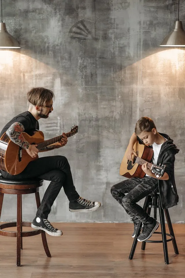 Two men sitting on stools, playing a classical guitar. The older one is teaching the younger one, a pre-teen, how to play guitar.