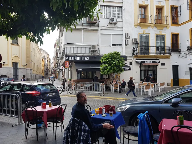 A man is sat at a small cafe table with a blue cloth in a street in Seville, Spain with typical Spanish buildings opposite