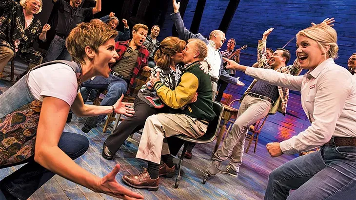 Two women frame a group of actors celebrating onstage as two characters kiss