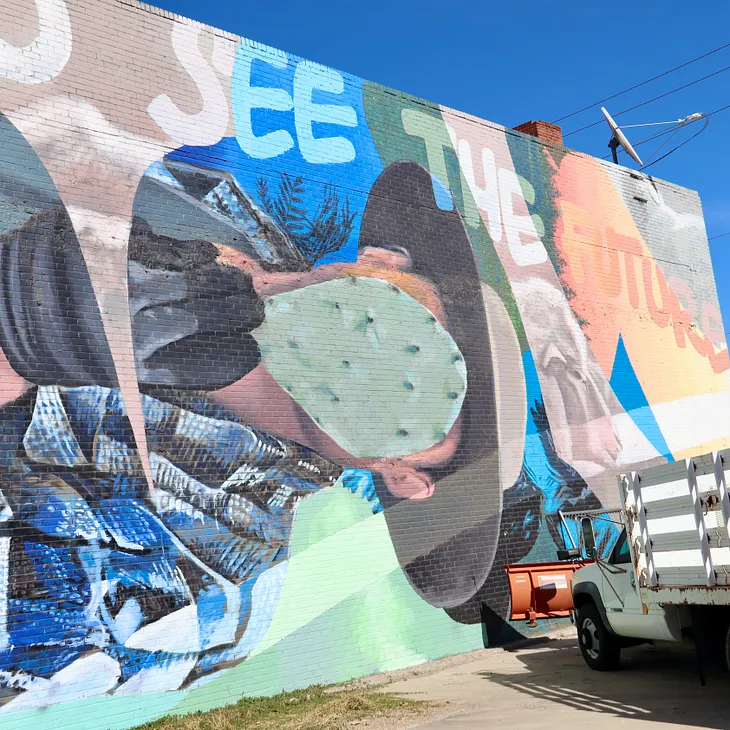 Murial, of a man holding a cactus leaf up to his face dressed like a field worker by the words “See The Future”, by a truck.