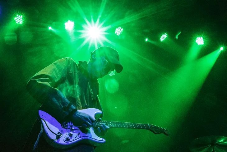 Cox in ballcap playing guitar under the glare of a green stagelight.