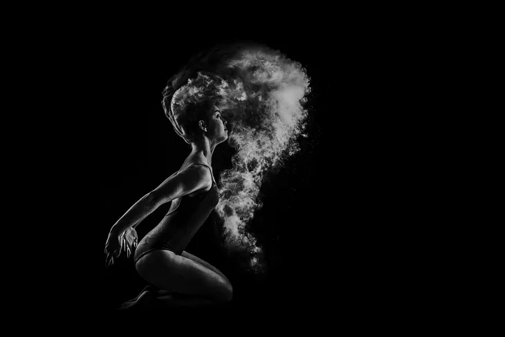 Black and white photo: female figure kneeling, a kind of fog or mist in front of her and hair in motion, wearing a swimsuit or a kind of leotard. (This is a much cooler photo than I’m making it sound. The figure is clearly in motion but I think that’s indicated by the appearance of the hair, and the mist or fog that looks displaced like something just moved through it.)