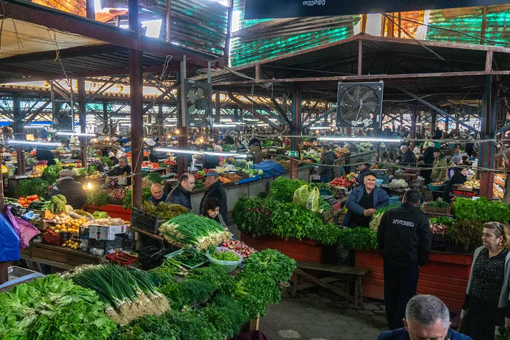 A view of a covered market hall in Kutaisi, Georgia. There are displays of fresh veggies along with vendors and shoppers.