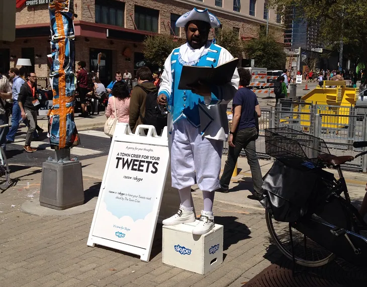 A man, dressed elaborately as an 18th century town crier, stands on a soap box next to a sign reading, “Town crier for your Tweets.”