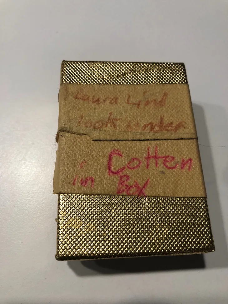 Shiny gold jewelry box, on the lid of which are two pieces of masking tape with these words in red ink: “Laura Lind look under Cotten in Box”