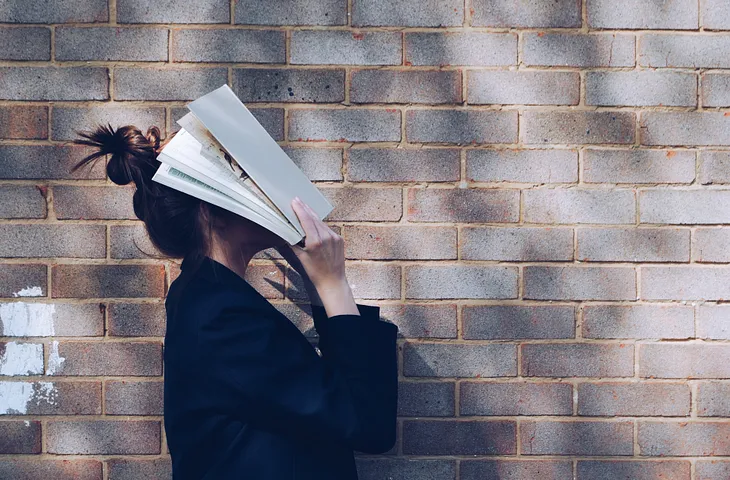 Person standing in front of a brick wall looking up and holding a book over their face.