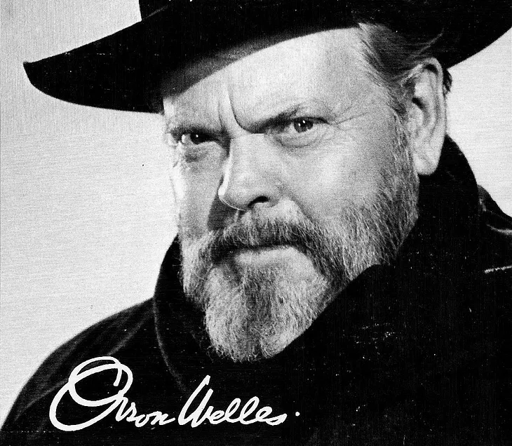 A black and white photograph of Orson Welles. He is wearing a thick black coat and scarf and a wide-brimmed black hat. He is older, late 50s, with a greying beard. He is looking directly at the camera with a smouldering expression. The photo is signed by the actor.