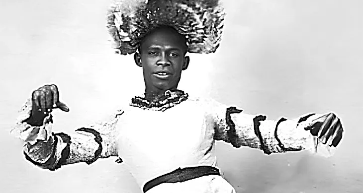 Black and white photograph of a Black Man in a white dress with dark ribbons on the sleeves and a fabulous headpiece that is really hard to describe.