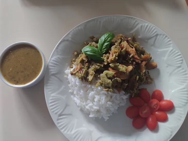 A white dish plated with white rice, green cooked karela aka bitter melon and decorated with cherry tomatoes, halved and arranged to replicate a red flower. On it’s left side a tiny bowl of green dahl soup a delicious addition to the meal.