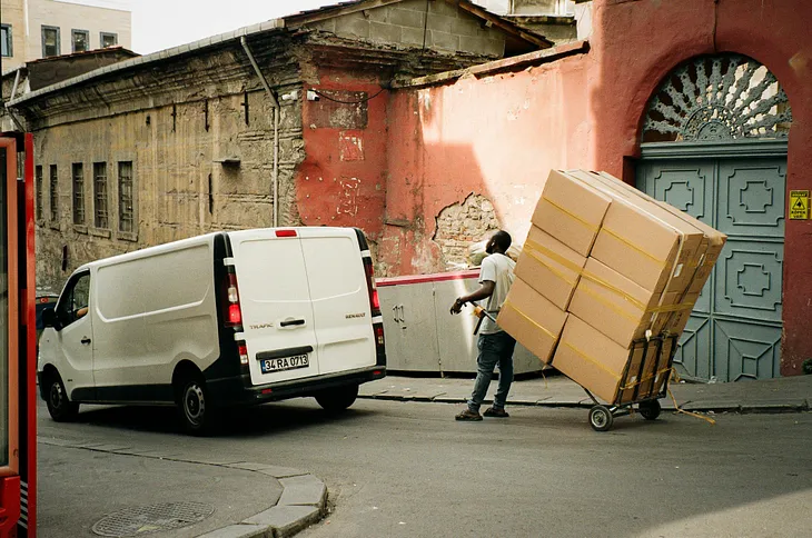 Delivery worker transporting large stack of cardboard boxes on a hand truck beside a white Renault van in an old city street with rustic buildings.
