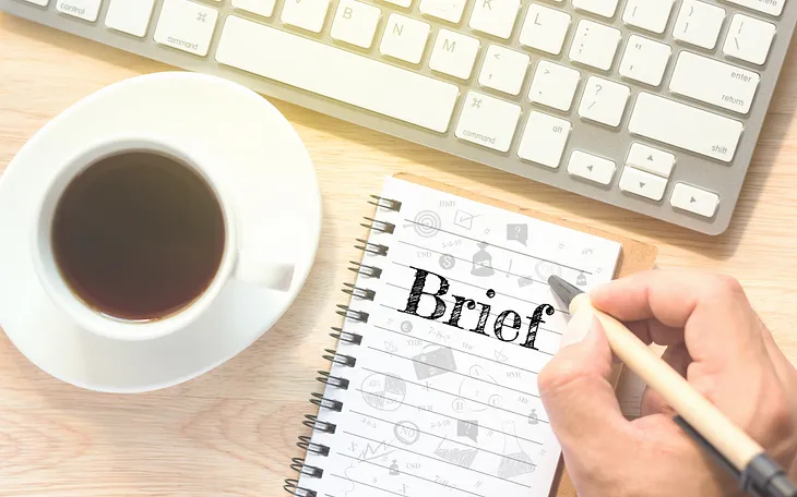 Picture of s hand writing “Brief” on a notepad next to a keyboard and mug of coffee