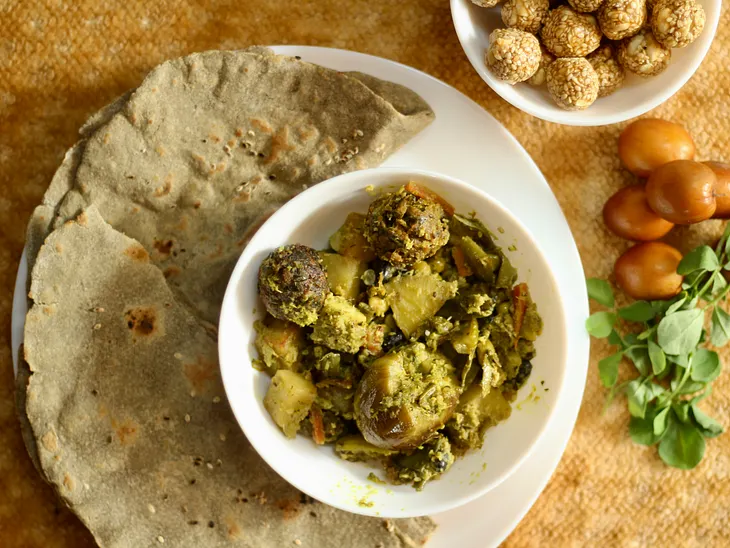 On the plate, a flatbread is served alongside a bowl of vegetables mixed in masala. Beside the plate, there’s another bowl with roundel sweets and a small fruit.