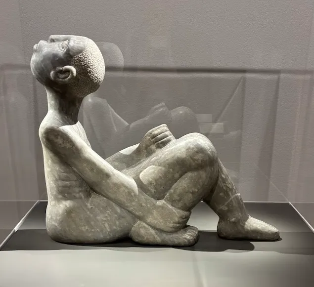 Sculpted in stone, a racially ambiguous male figure sits with his head twisted an impossible 180 degrees, aimed upward at the sky.