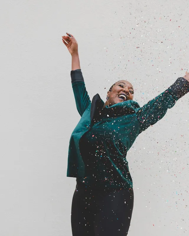 Vibrant young black woman in turquoise jacket with arms raised and light confetti coming down on her.