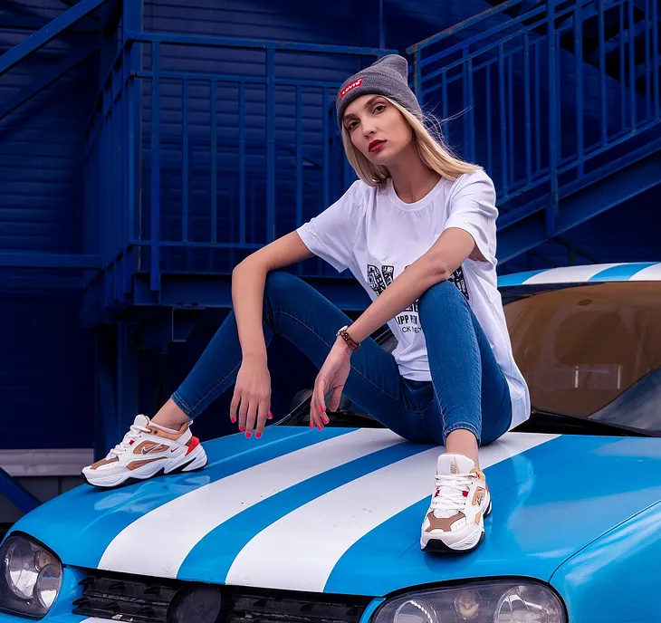 young woman sits on car, long blond hair, beanie, wears t-shirt, jeans, trainer and beanie, car has a white stripe on bonnet and roof