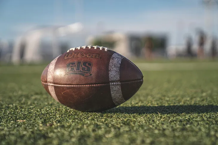 A football sits alone on a green turf football field.