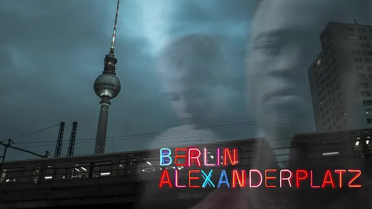 Poster for Berlin Alexanderplatz. It is a photo of a train passing by a spire at night. There are dark clouds in the sky. There are transparent images of Mieze and Francis transposed on top of it with the words Berlin Alexanderplatz in neon colors and as neon signage in the bottom right corner.