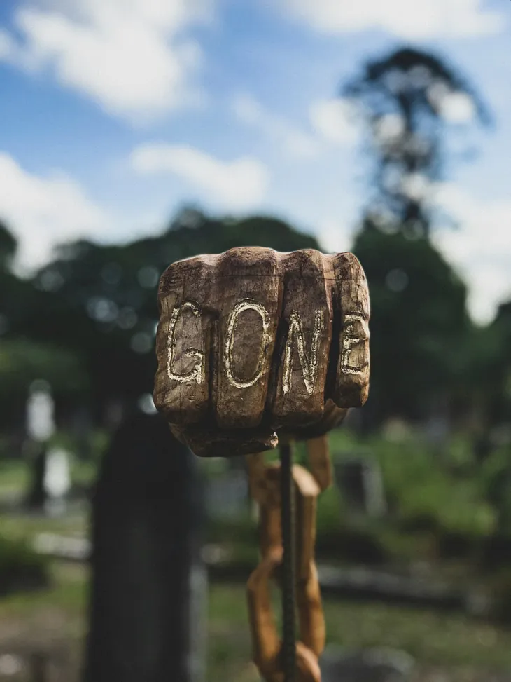 A wooden sign engraved with the word “GONE,” set against a blurred cemetery background.