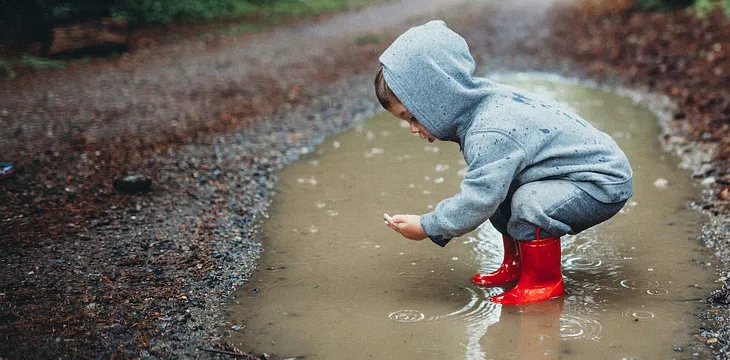 A toddler studying something that it has picked up out of the puddle they are standing in