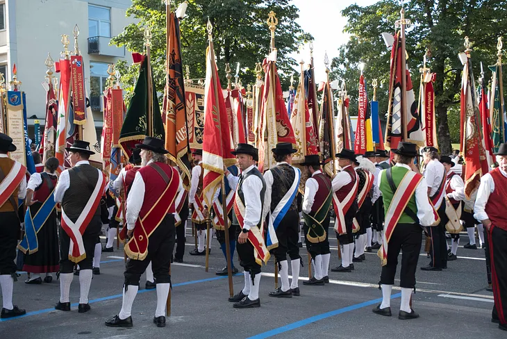 Austrian men wearding lederhosen standing on the road and holding different type of flags that represent thier villages or town.