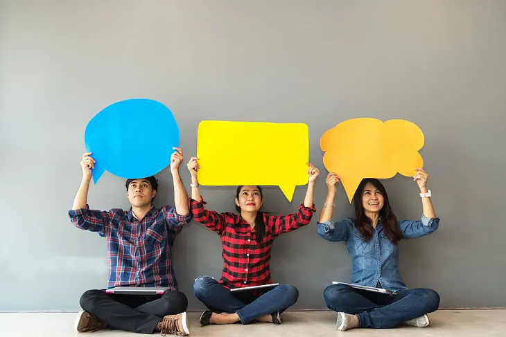 A man and two women sit on the floor holding cut out speech bubbles over their head.