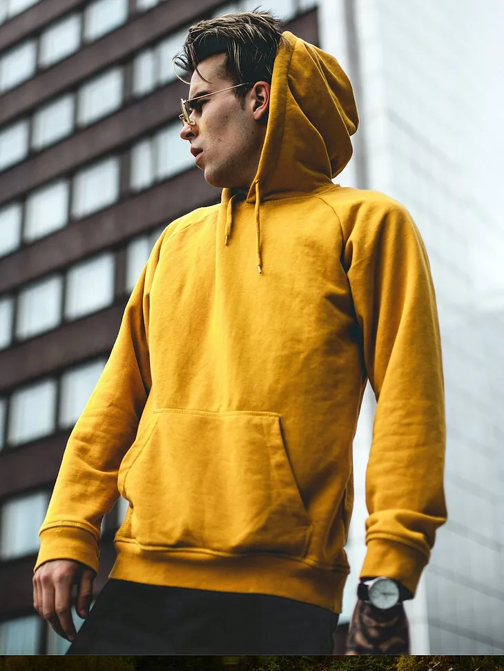 Young man in the city, looking over to his right, `wearing a mustard color pullover hoodie with kangaroo pockets