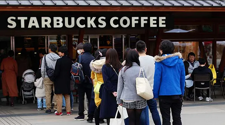 A photo of people waiting in line at a coffee shop. The first person in line is placing their order (the message being sent), the barista is processing the order for the person at the front (the message being received), and everyone else is waiting their turn (other messages waiting to be processed). This image emphasizes the First-In-First-Out (FIFO) nature of message queues, where messages are processed in the order they are received.