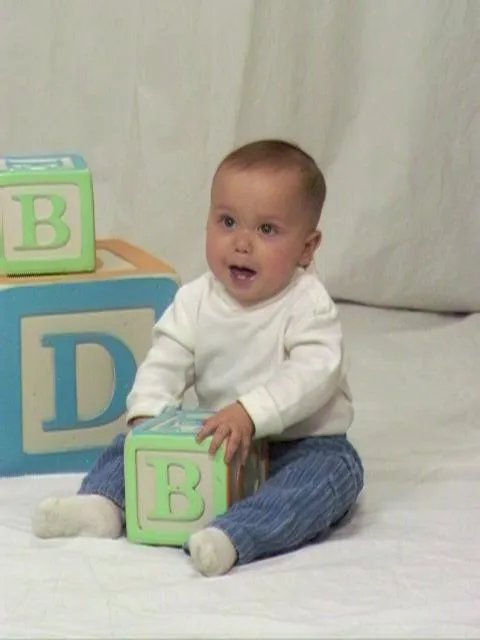 Picture of an 8 month old boy with a white turtleneck and light blue corduroy pants, holding an oversized block.