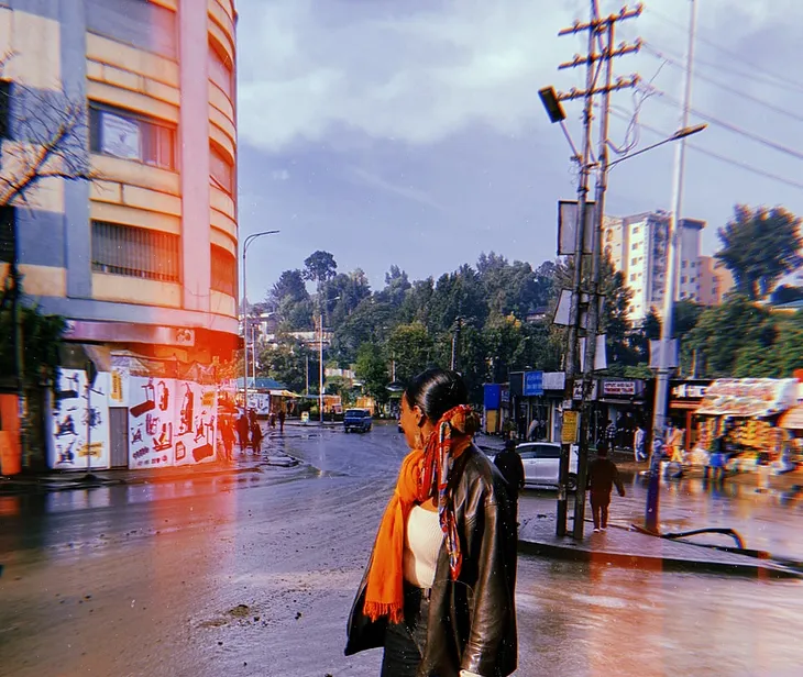 A person wearing a bright orange scarf standing in Piassa Town during kiremt season with their face turned away from the Camera