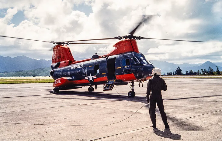A photograph of a twin-rotor military helicopter on the ground with rotors turning. In the foreground is the crew chief, in flight suit and helmet, watching the aircraft.
