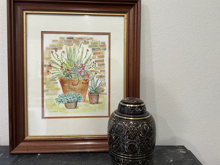 A large framed watercolor of 3 succulents and a black and gold urn sit on a gray side table