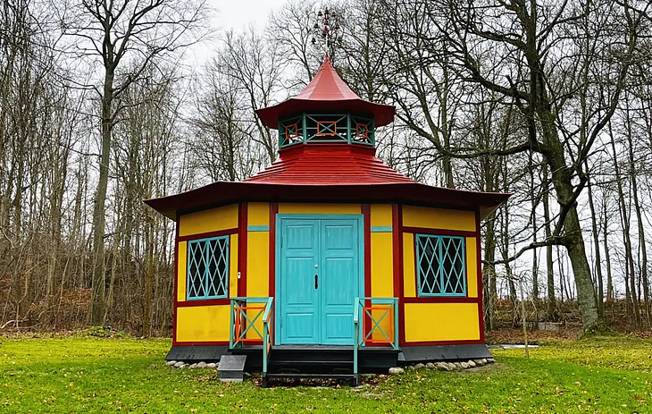 A small, hexagonal, 18th-century teahouse, painted bright yellow. with red and turquoise trim. Surrounded by bare wintery trees and set in a bright moss-green lawn.