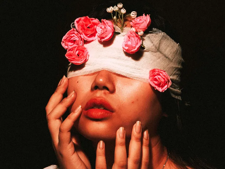 Portrait of a woman with a bandage covering her eyes, adorned with bright pink roses against a dark background, with her hands gently touching her face.