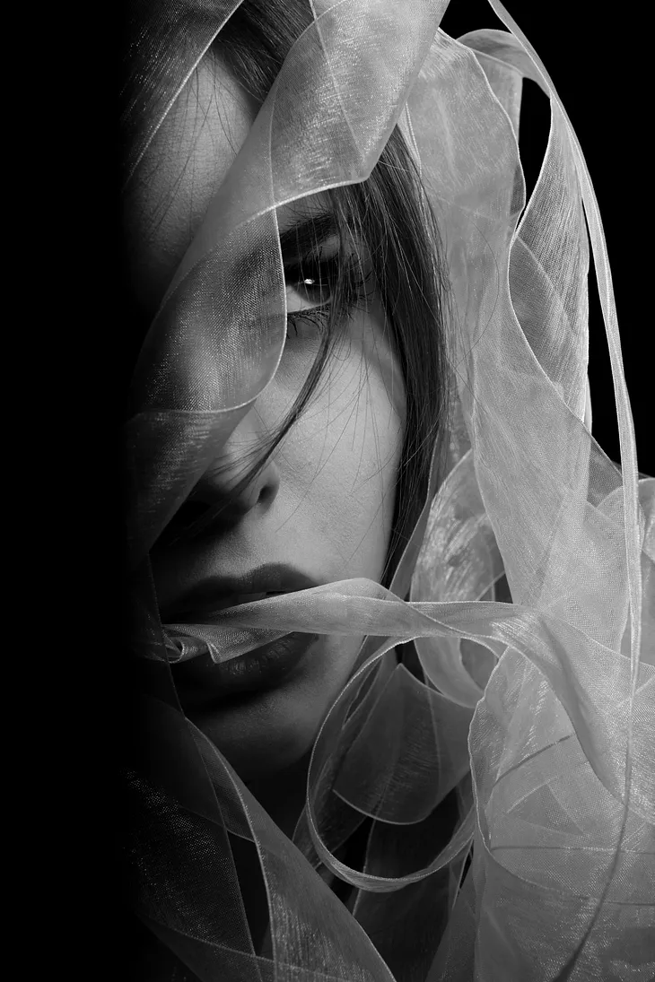 Black and white close-up portrait of a woman’s face partially obscured by sheer, textured fabric, highlighting her intense gaze and the delicate details of the material.