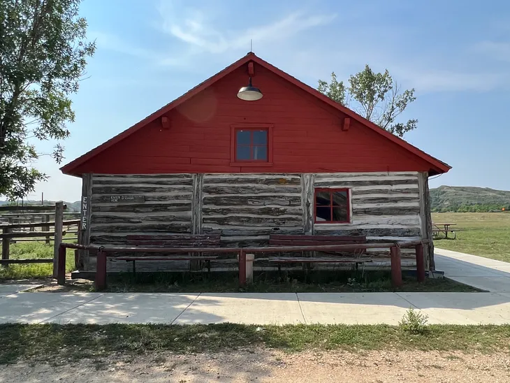 This is a photo of a log cabin with a red roof surrounded by beautiful countryside, green grass, and mountains in the background.