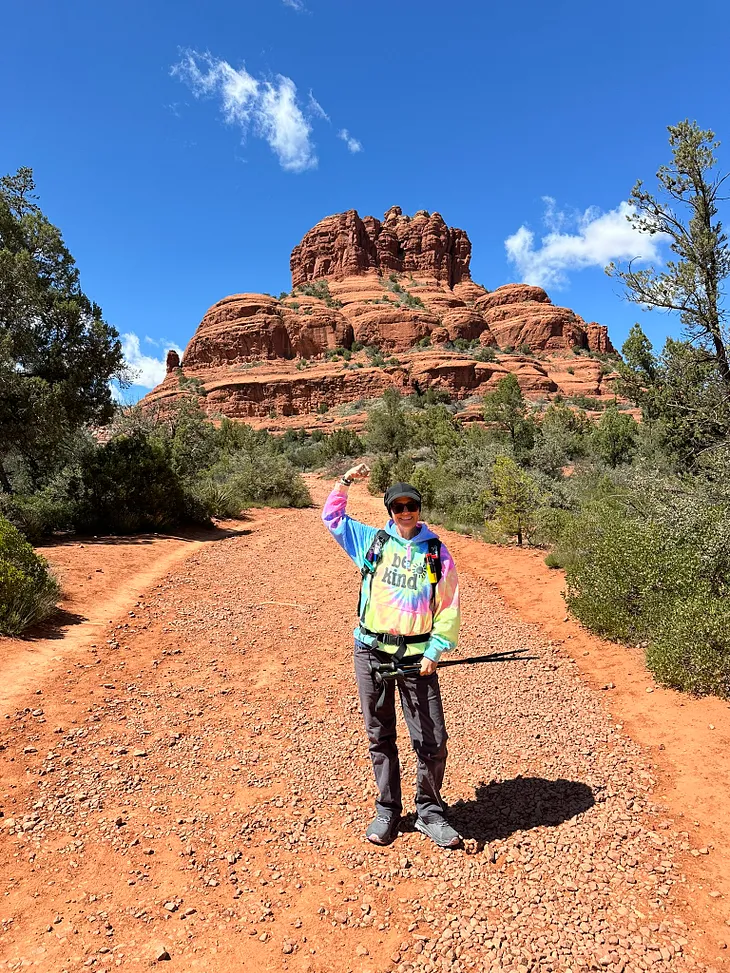 CC in a rainbow hoodie standing in front of Bell Rock red rock vortex in Sedona with a strong arm up