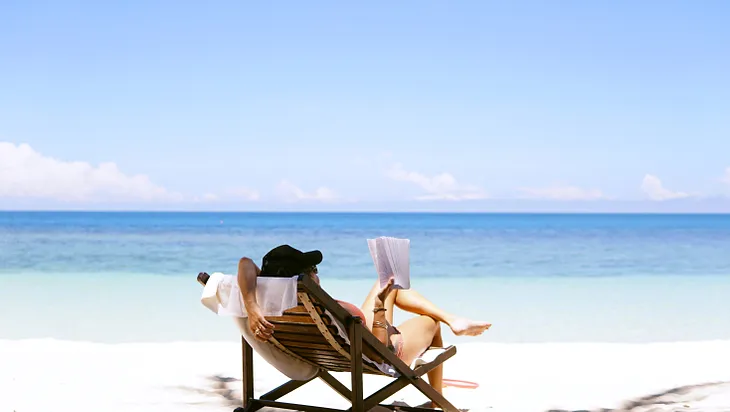 A woman sits on a brown wooden beach chair with a book