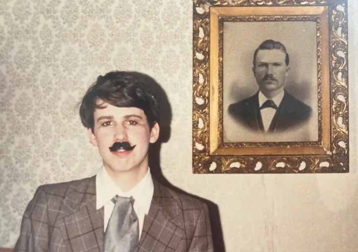 A young teen in theater makeup wears a period suit and wears a fake mustache while standing before a black and white portrait of a man.