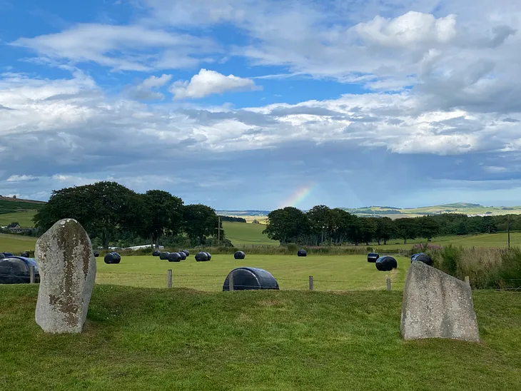 View of a partial rainbow disappearing into clouds, from within an ancient stone circle, in the middle of the countryside.
