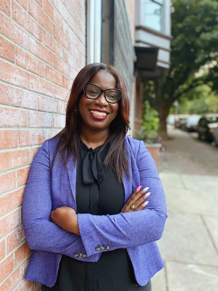 This is a headshot of Atiyah. She is wearing a purple blazer with a black shirt that has a bow in the front. Her nails are painted with pink designs. She is wearing red lipstick and she is smiling. Atiyah is also wearing black glasses. Her hair is brown and it is straight with side bangs.