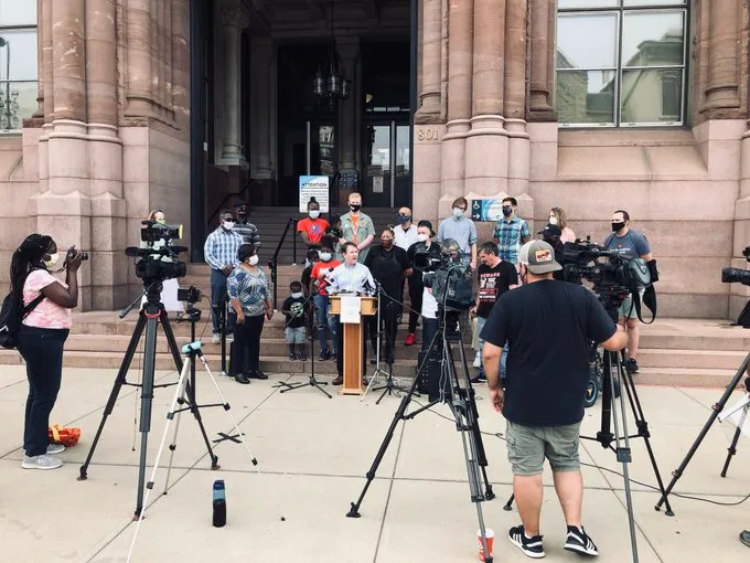 Jack Jose stands at the podium on the steps of Cincinnati City Hall, surrounded by Neighborhoods United supporters and media.