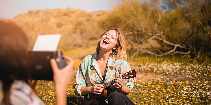 A woman is sitting in a field of wildflowers, playing a ukulele and laughing joyfully while her friend takes a photo of her. The sun is shining brightly, and the background shows bushes and a clear sky, creating a vibrant and cheerful scene of friends capturing a moment of happiness in nature.