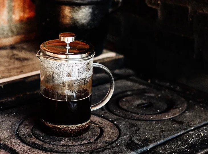 A French press coffee half-full of coffee sits on top of an old stove.