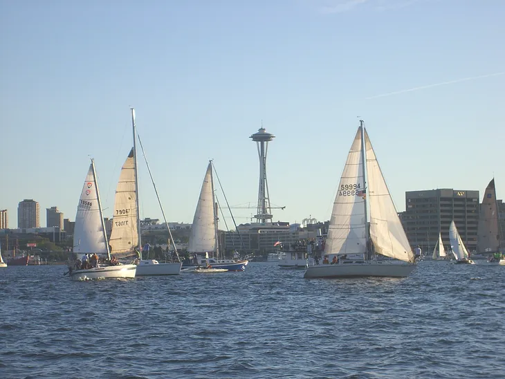 Picture taken from Lake Union of sailboats framing Seattle Space Needle. Choppy water in foreground, blue sky in background.