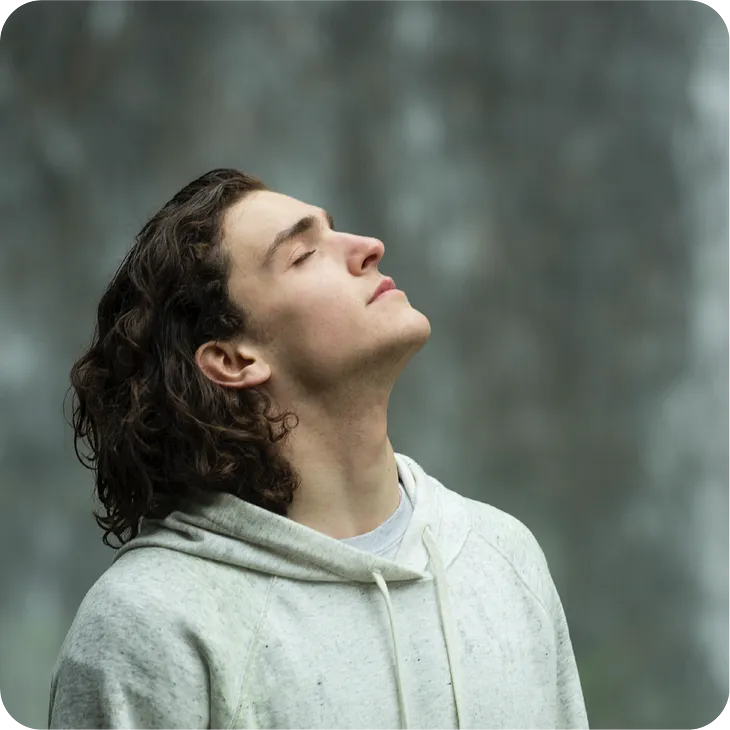 Man with long hairs in gray pullover hoodie looking up at sky with eyes closed with a sense of acceptance and relief.