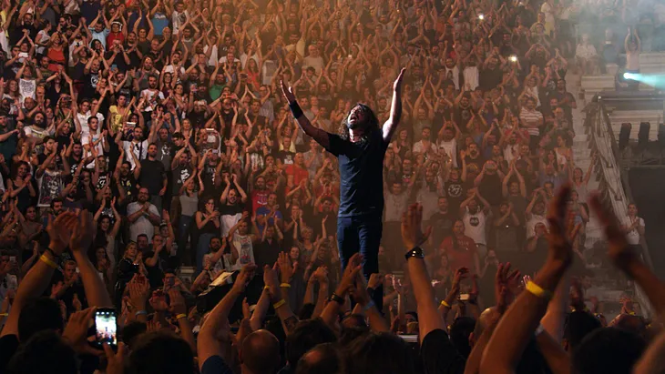 Dave Grohl, Foo Fighters frontman, arms up, surrounded by a huge audience, in a big stadium concert. 1000 True Fans Theory.