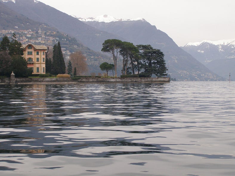 A lake is surrounded by mountains. A town is visible in the distance rising up from the lake. There is a peninsula with a building and trees in the foreground.